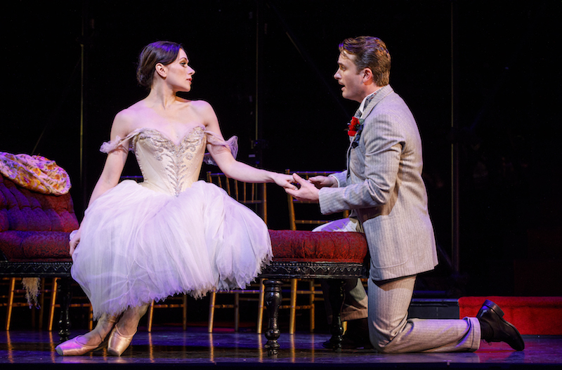 Dvorovenko in a a pale pink tutu and off the shoulder bodice and pointe shoes sits on a chair. A man in a suit kneels singing to her and holding her hand.